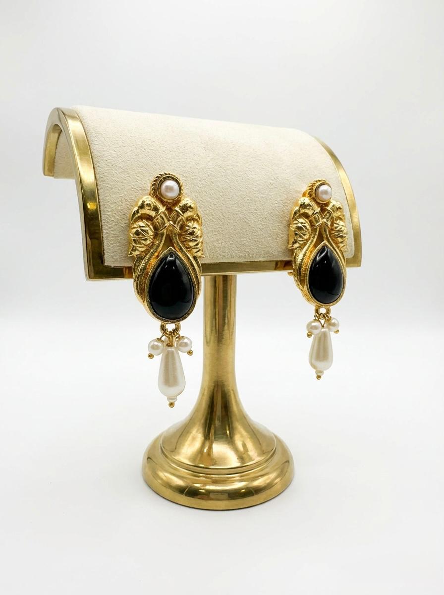 A pair of sophisticated black jhumka-style drop earrings resting on a jewelry stand, featuring a dark cabochon stone set in an ornate brass frame with classic pearl embellishments.
