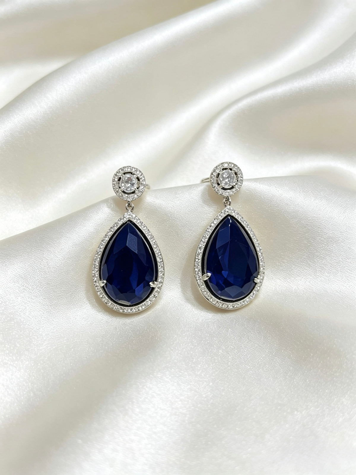 A pair of teardrop-shaped drop earrings with deep blue gemstones surrounded by small clear stones in silver-toned settings, displayed on draped white satin fabric.