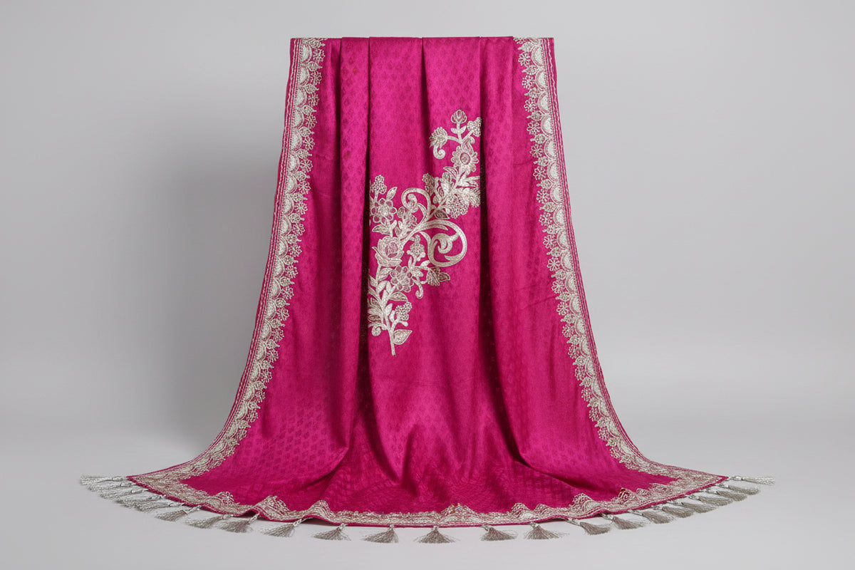 A pink Kashmiri Aari embroidered marina scarf with tassels hanging at the ends, displayed on a wooden hanger with greenery in the background.