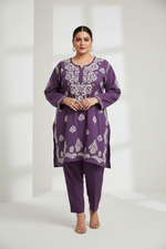 A woman modeling a purple kurta set with white floral embroidery, paired with matching purple pants, in a bright, minimalist white studio setting.