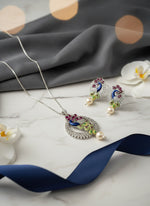 A rhodium-plated brass pendant with a peacock design accompanied by earrings, featuring vibrant green, blue, enamel accents, as well as sparkling crystal white and ruby pink embellishments.