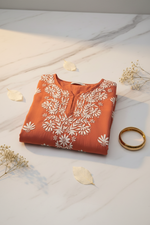 A rust-orange long-sleeve tunic top with white floral and leaf patterns, featuring a split neckline with button details, shown against a plain white background.