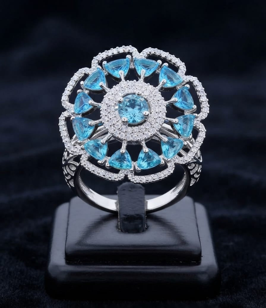 A silver-tone ring with a floral mandala design, featuring light blue gemstones and small clear accent stones, displayed on a black stand against a dark velvet background.