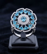 A silver-tone ring with a floral mandala design, featuring light blue gemstones and small clear accent stones, displayed on a black stand against a dark velvet background.