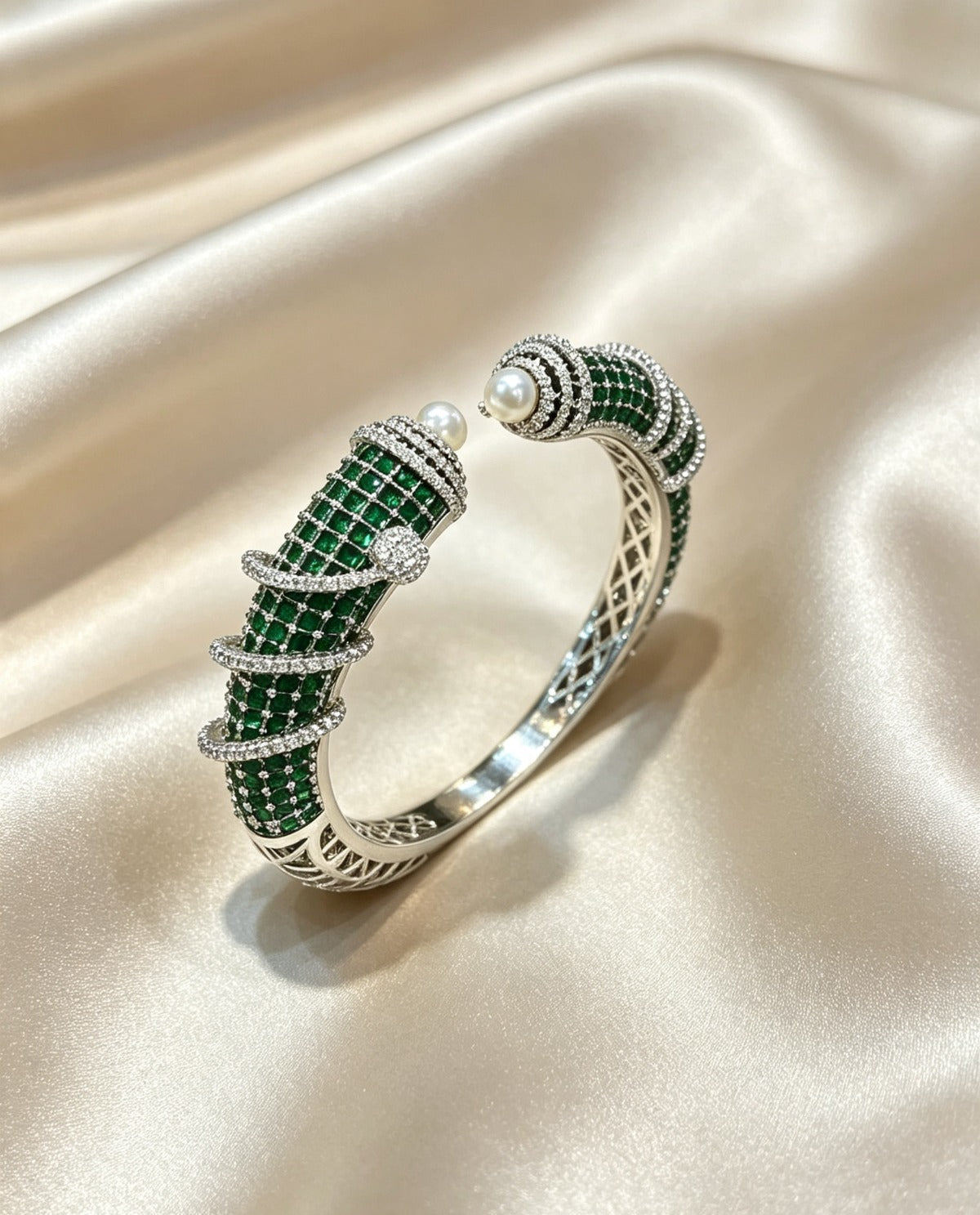 A silver-toned bangle bracelet featuring intricate green stone pavé sections, a single white pearl, and small clear stones, displayed on a draped beige satin fabric.