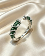 A silver-toned bangle bracelet featuring intricate green stone pavé sections, a single white pearl, and small clear stones, displayed on a draped beige satin fabric.