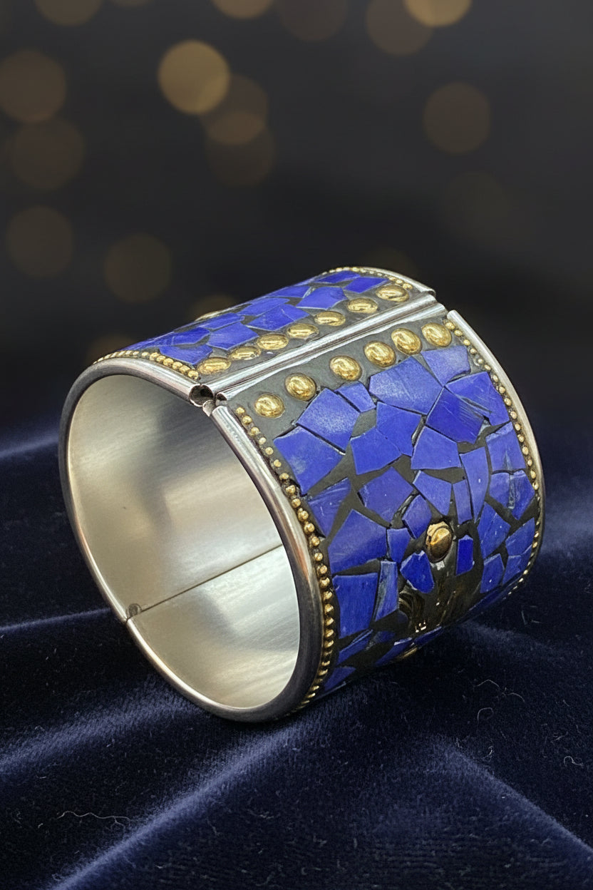 A silver-toned cuff bracelet featuring a mosaic inlay of blue lapis lazuli stones and small gold-tone studs and beaded borders, displayed on a dark textured background.