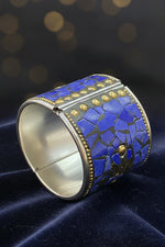 A silver-toned cuff bracelet featuring a mosaic inlay of blue lapis lazuli stones and small gold-tone studs and beaded borders, displayed on a dark textured background.