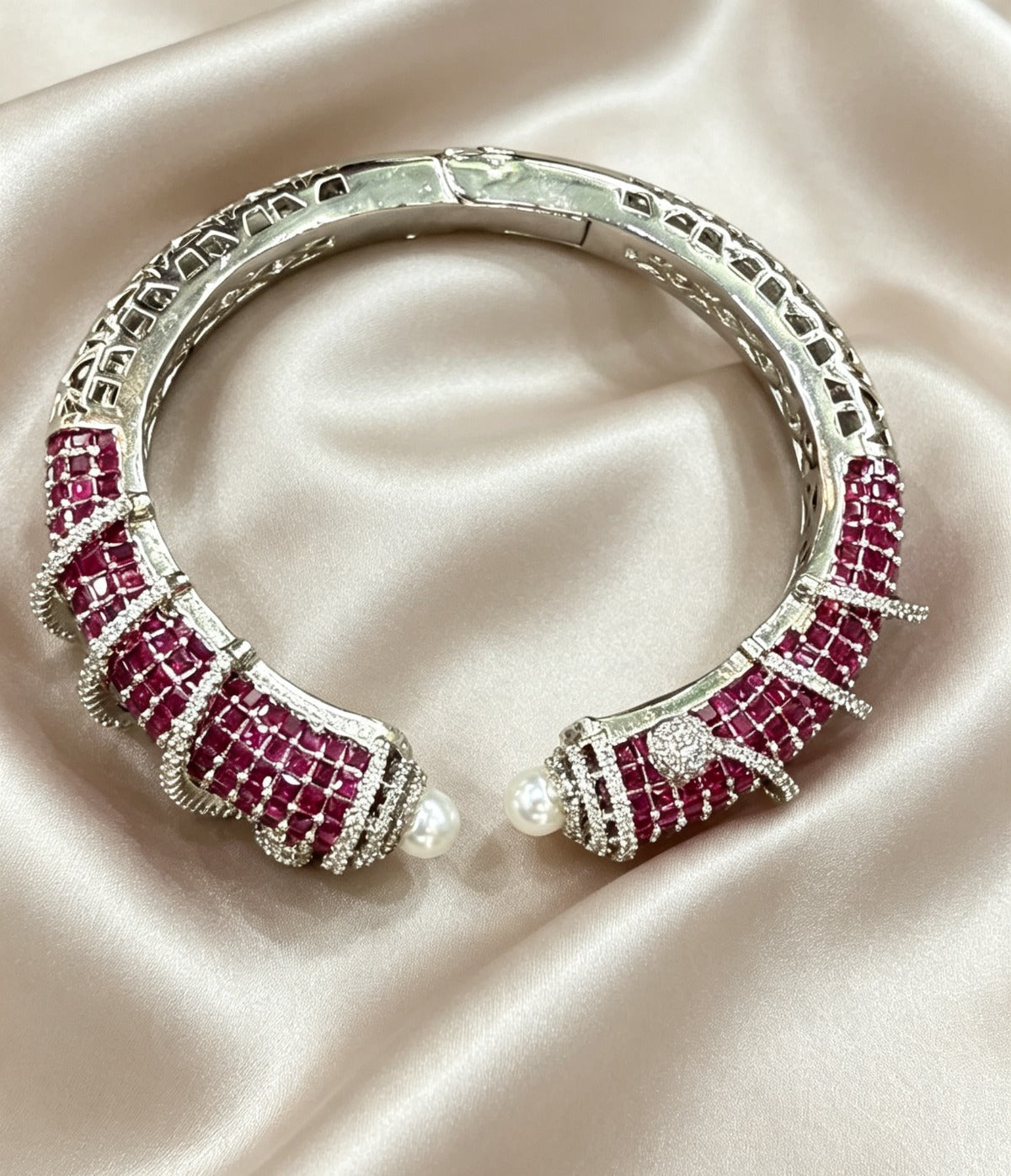 A silver-toned cuff bracelet featuring rows of deep pinkish-red round gemstones and small white pearl accents, displayed on draped champagne satin fabric.