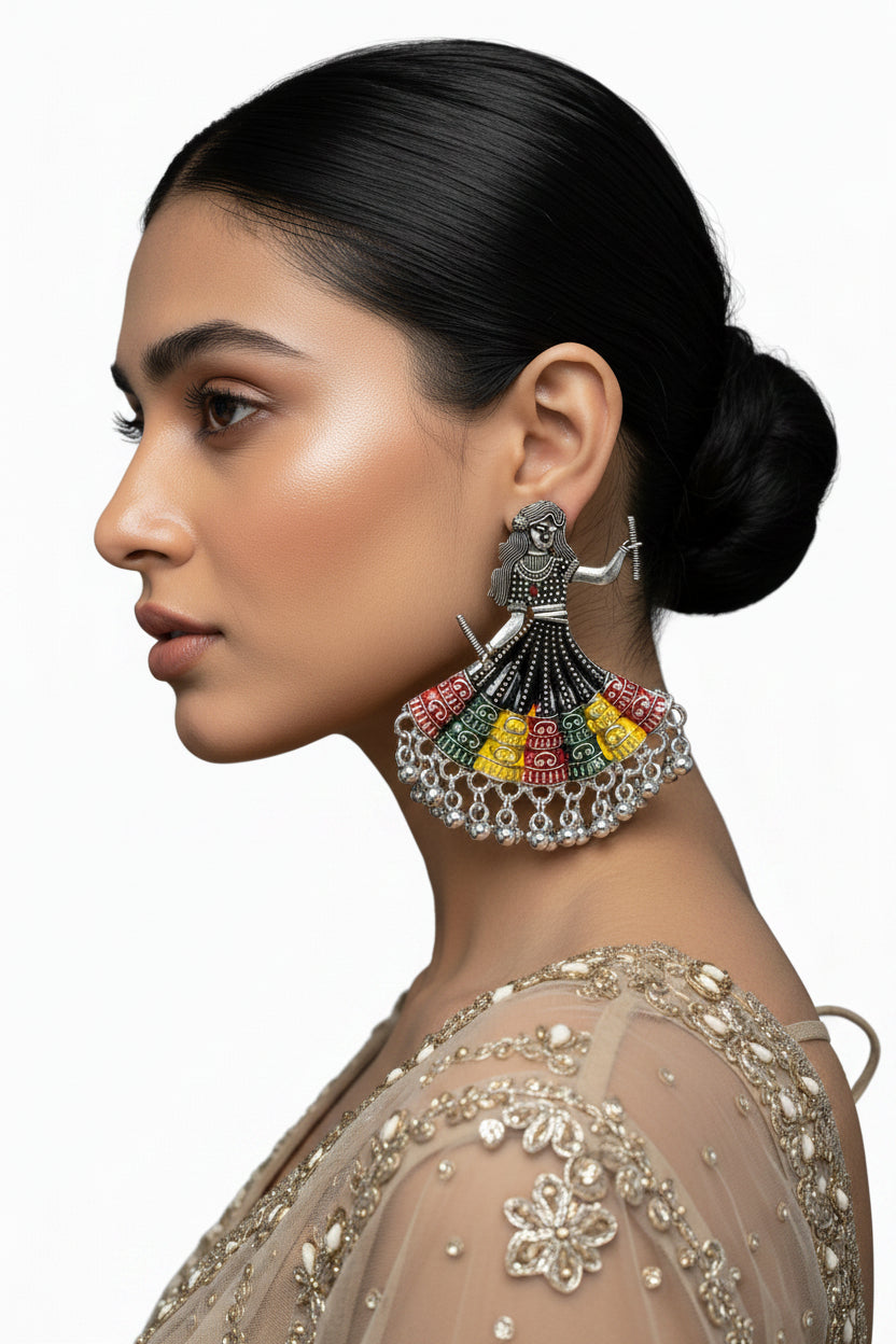 A silver-toned earring shaped like dancing women in traditional navratri chaniya choli dress, decorated with black beads and colourful enamel details in red, yellow, green, and blue, displayed on a white marble surface.