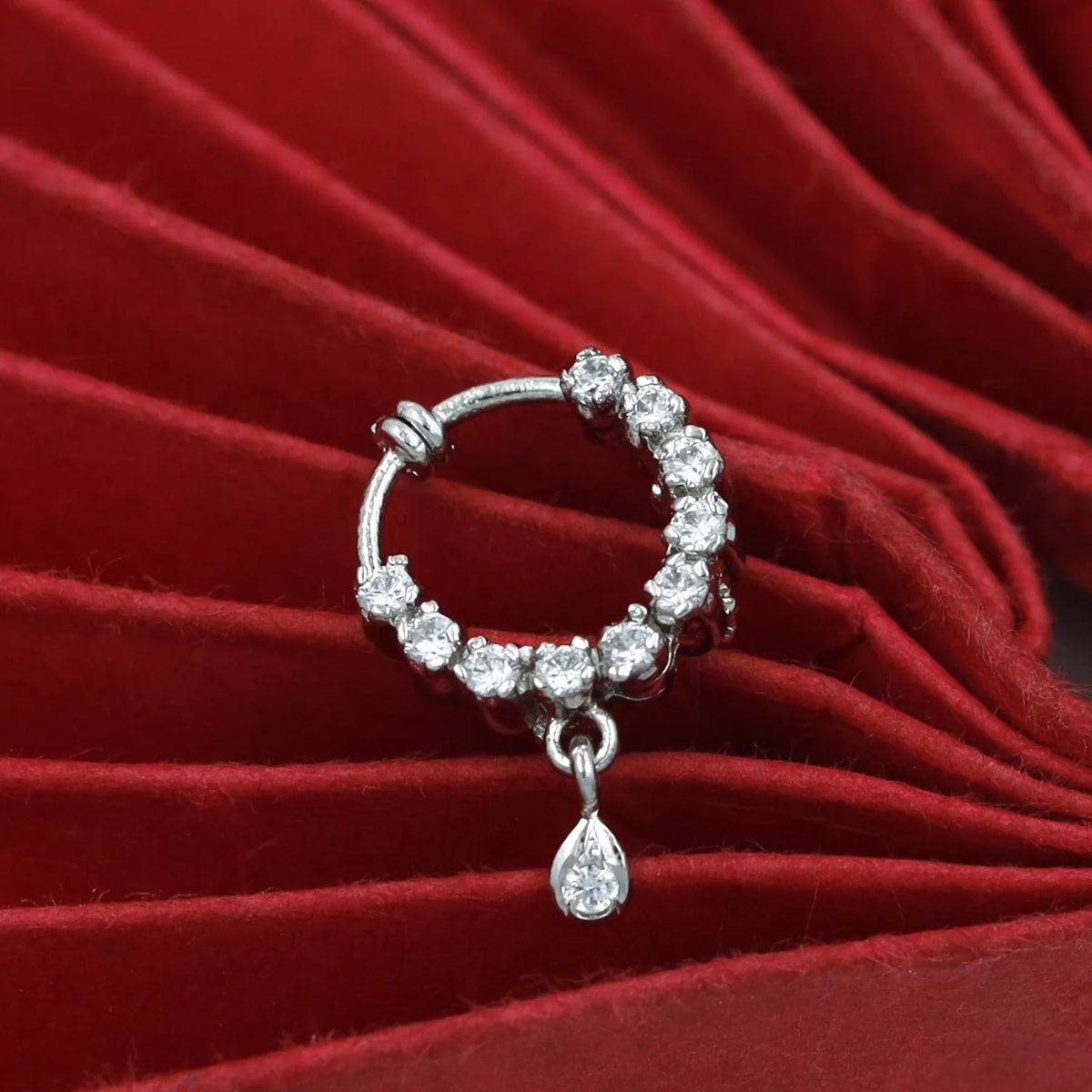 A silver-toned huggie hoop earring embellished with clear round stones and a dangling teardrop-shaped clear stone, displayed on a red velvet surface.