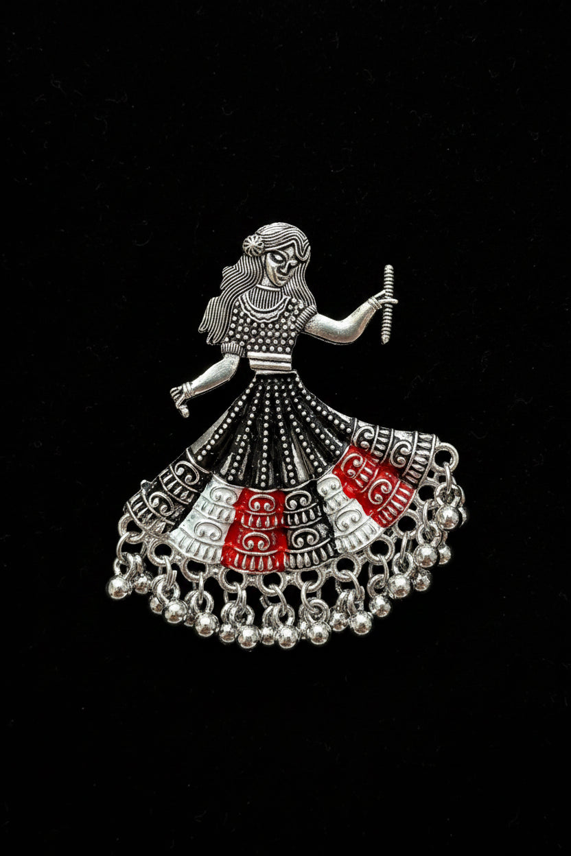 A silver-toned metal earring brooch depicting a traditional Indian folk dancer with a flared skirt patterned in black, white, and red, resting on a white marble surface.