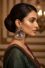A woman in profile wearing a large silver-toned traditional jhumka earring with red, white, and green stone accents, photographed in an elegant indoor setting with warm blurred lights in the background.