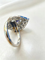 A silver-toned ring with a geometric, leaf-like pavé setting and rectangular blue gemstones, photographed on a soft white fabric background.