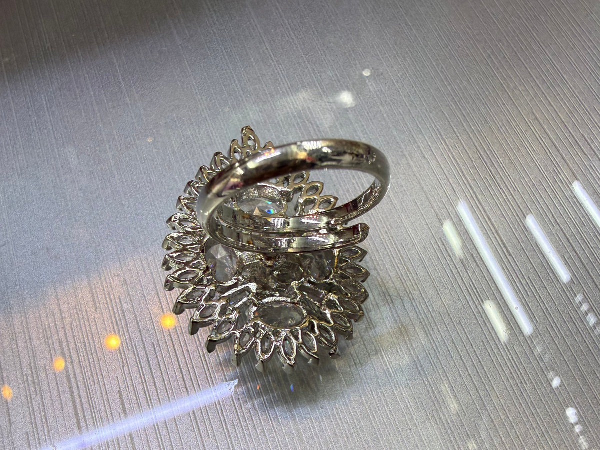 A silver-toned ring with a highly textured, intricate floral or sunburst-like pavé setting and a smooth band, resting on a reflective metallic surface.
