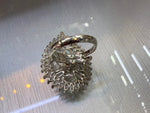 A silver-toned ring with a highly textured, intricate floral or sunburst-like pavé setting and a smooth band, resting on a reflective metallic surface.