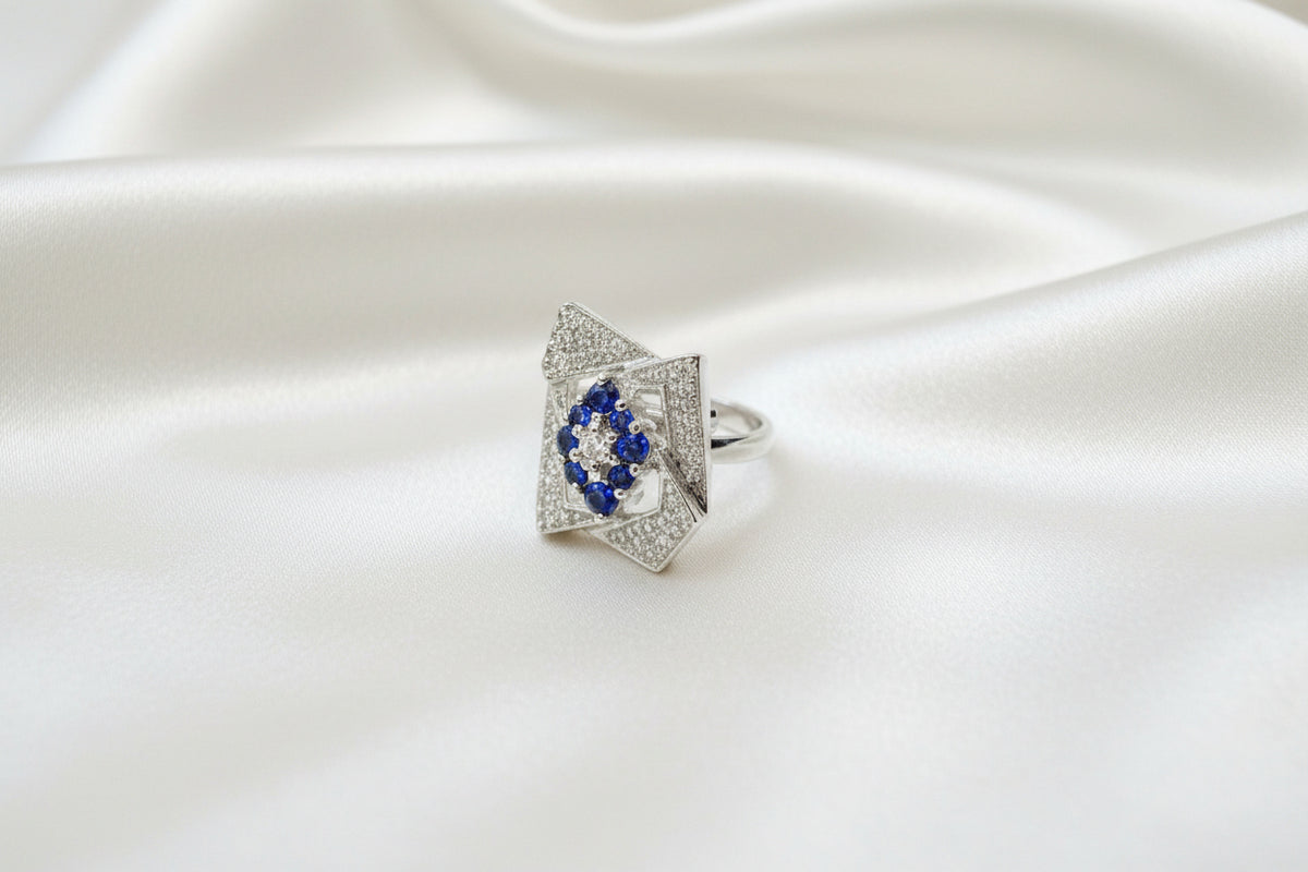 A silver-toned ring with a rectangular pavé setting and small blue gemstones, photographed on a textured blue fabric background.