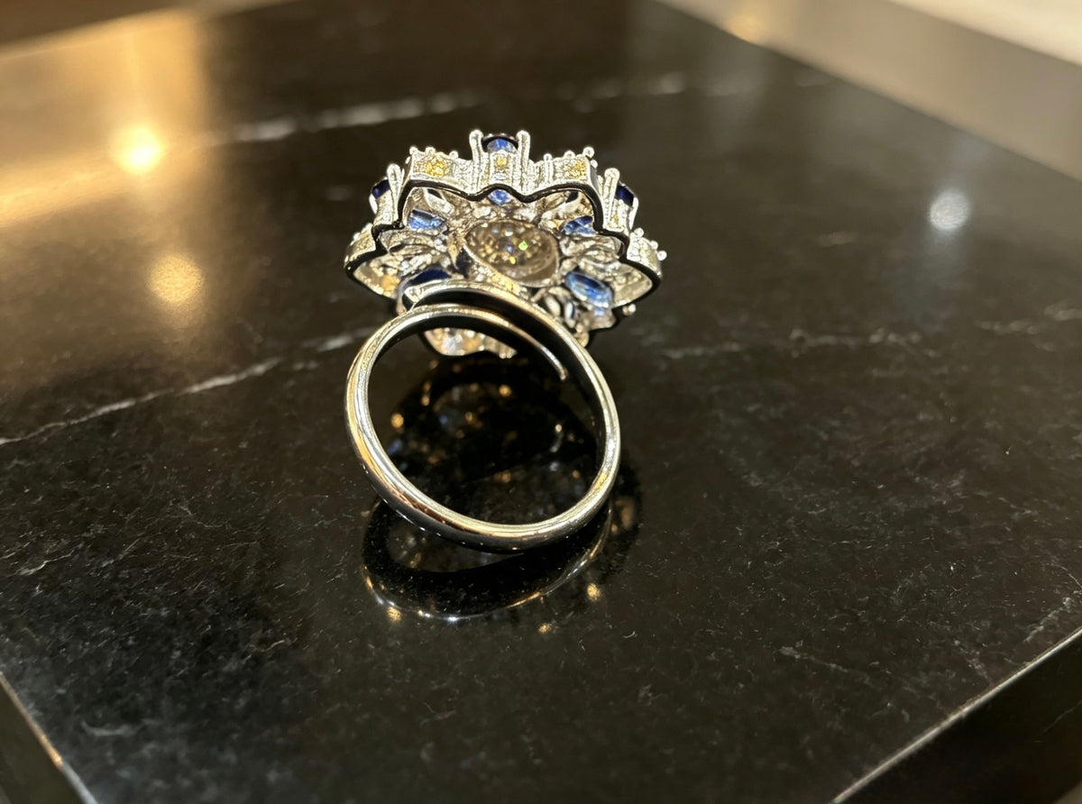 A silver-toned ring with an ornate floral or crown-like setting featuring blue and clear stones, displayed on a glossy black marble surface.