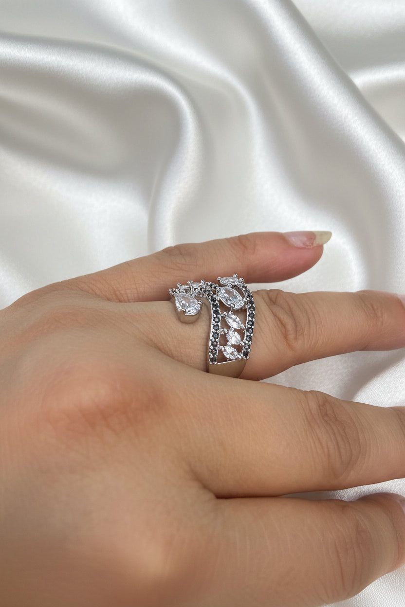 A silver-toned ring with clear gemstones worn on a person's finger against a black background.