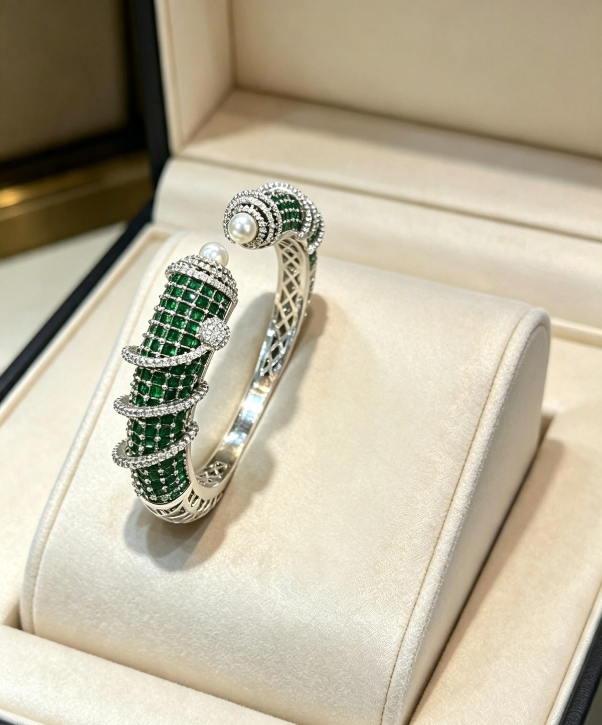 A silver-toned twisted bangle bracelet with a row of green stones and small clear stones, displayed on a cream jewelry box cushion.