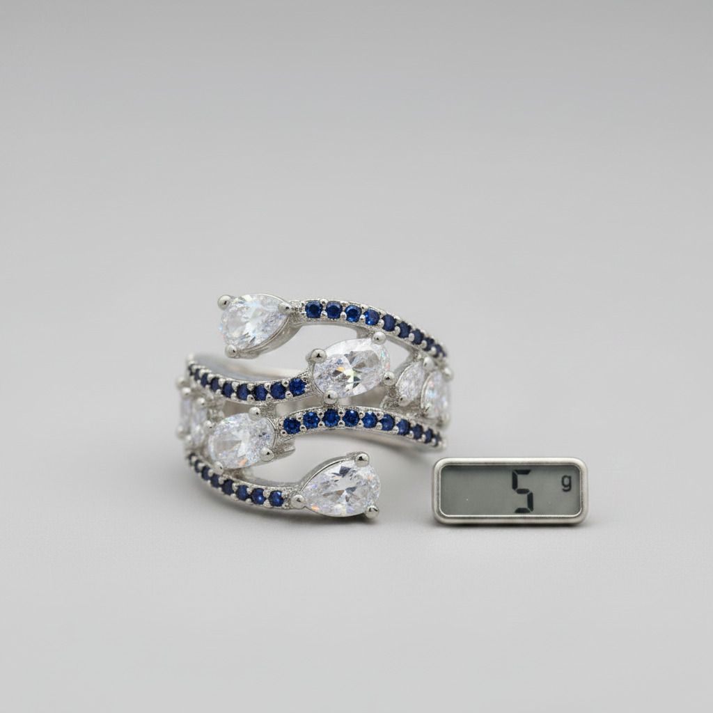 A silver-toned wrap bracelet with pear-cut clear stones and rows of deep blue round stones, shown on a light gray background next to a small digital scale displaying 5 g.