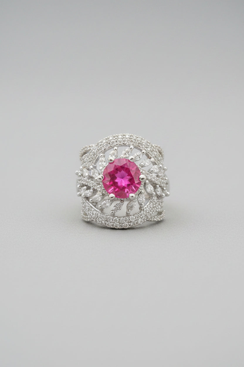 A silver ring with a round pink gemstone and pavé-set clear stones, held between fingers against a dark textured background.