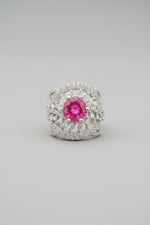 A silver ring with a round pink gemstone and pavé-set clear stones, held between fingers against a dark textured background.