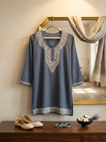 A slate blue-grey tunic with intricate white embroidery on the neckline and hem, hanging on a gold hanger on a wooden shelf. The scene is styled with a matching scarf draped over a gold mirror, beige flat shoes, and a small bowl of decorative items on a wooden table.