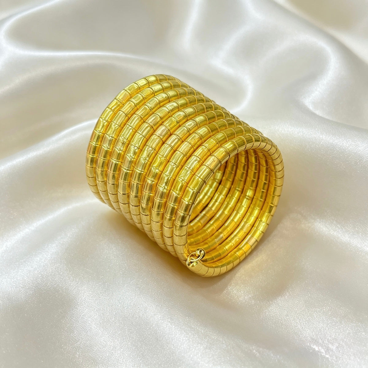 A stack of wide, textured gold bangles arranged in a cylinder on a draped white satin fabric background.