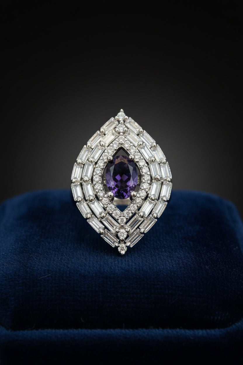 A teardrop-shaped silver finger ring featuring a central oval deep purple gemstone surrounded by multiple rows of baguette and round clear stones, displayed on a textured blue velvet surface.
