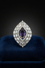A teardrop-shaped silver finger ring featuring a central oval deep purple gemstone surrounded by multiple rows of baguette and round clear stones, displayed on a textured blue velvet surface.
