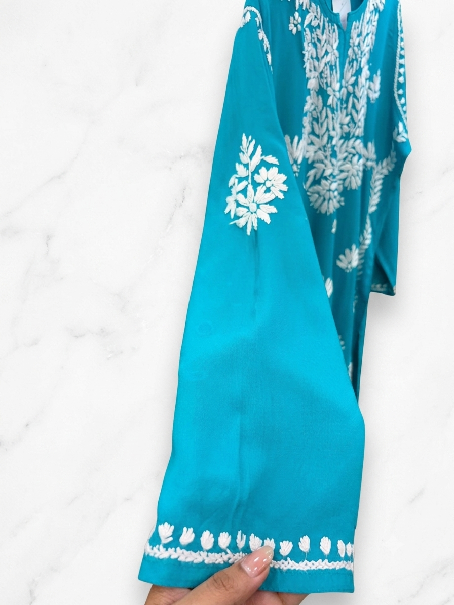A turquoise blue long-sleeve tunic or kurta with white floral embroidery and a matching embroidered border on the sleeve, held by a hand against a white marble background.
