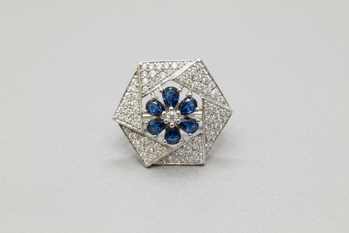 A vintage-style hexagonal brooch featuring a silver-toned pavé setting with a central blue floral motif, resting on a textured blue velvet surface.