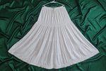 A white tiered skirt with embroidered details and a ruffled hem, hanging on a hanger against a neutral background.