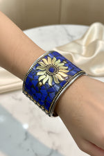 A wide silver-toned cuff bracelet with a mosaic blue and gold floral design, worn on a person's wrist.