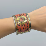 A wide silver-toned cuff bracelet with a mosaic design, featuring terracotta red tiles, gold-toned leaf shapes, and a central floral motif, shown worn on a wrist.