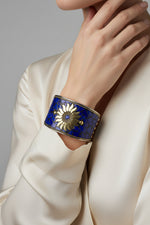A wide silver-toned cuff bracelet with a mosaic of deep blue glass tiles and a raised gold-tone sunflower motif on the front, shown worn on a person's wrist.