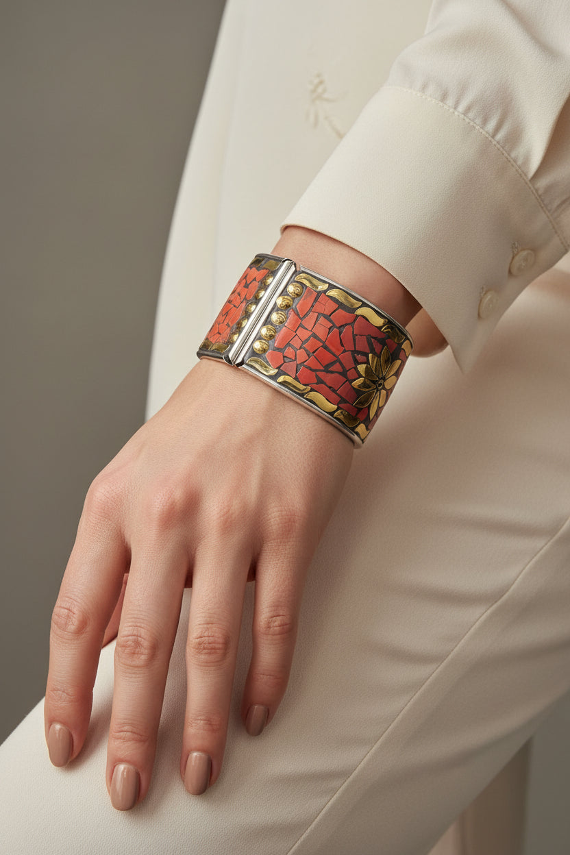 A wide silver cuff bracelet with a textured red-orange mosaic inlay and small gold decorative accents, displayed on a black surface.