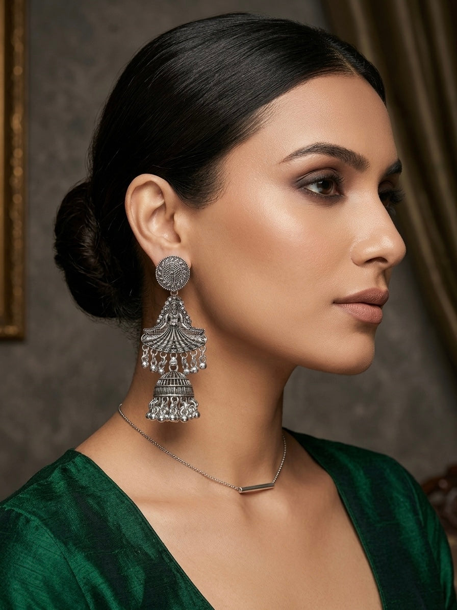 A woman in profile wearing a large silver-toned jhumka earring and a delicate silver necklace, dressed in a deep green velvet top against a dark, elegant indoor background.