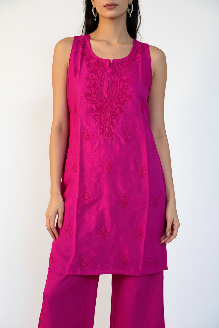 A woman modeling a bright magenta pink kurta set with matching trousers, featuring embroidered detailing on the chest, set against a textured white curtain backdrop.
