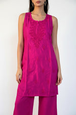 A woman modeling a bright magenta pink kurta set with matching trousers, featuring embroidered detailing on the chest, set against a textured white curtain backdrop.