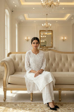 A woman modeling a white embroidered kurta and matching trousers in a bright, elegant indoor setting with soft sunlight and cream-colored walls.