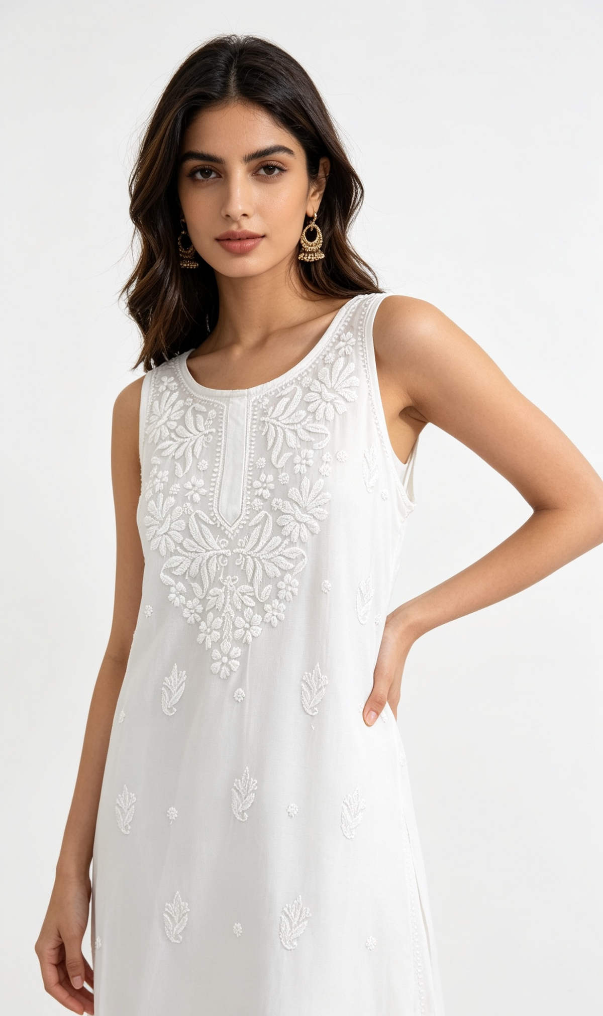 A woman modelling a sleeveless white dress with intricate floral embroidery on the bodice and scattered embroidered motifs down the skirt, set against a plain white studio background.