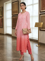 A woman models a light pink embroidered kurta and matching trousers set, holding a beige clutch bag in a bright indoor gallery setting.