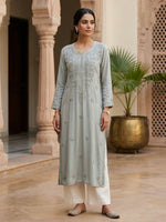 A woman models a light sage green embroidered kurta with white pants in an ornate indoor courtyard setting.