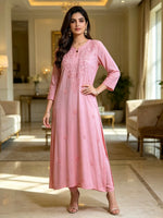 A woman models a long pink embroidered kurta in a bright, elegant living room. The kurta features a round neckline, three-quarter sleeves with embroidered cuffs, and delicate floral embroidery on the front and scattered down the skirt.
