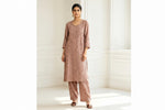 A woman models a three-piece dusty rose pink ethnic kurta set with matching palazzo pants. The set features intricate floral embroidery and is styled with gold earrings and sandals in a bright, white-paneled room.