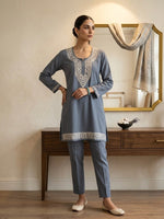 A woman models a two-piece traditional outfit consisting of a dusty blue-gray kurta with intricate white embroidery around the neckline and hem, paired with matching slim-fit trousers. She is standing in a styled room with a wooden console table and a mirror in the background.