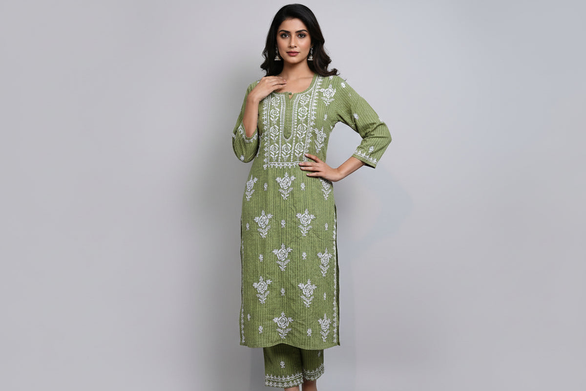 A woman standing on a sidewalk with wearing a green pink colour traditional Lucknowi chikankari kurti with floral patterns and palazzo pants, holding a mobile phone and a bag.