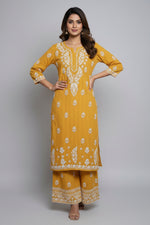 A woman standing on a sidewalk with wearing a mustarad colour traditional Lucknowi chikankari kurti with floral patterns and palazzo pants, holding a mobile phone and a bag.